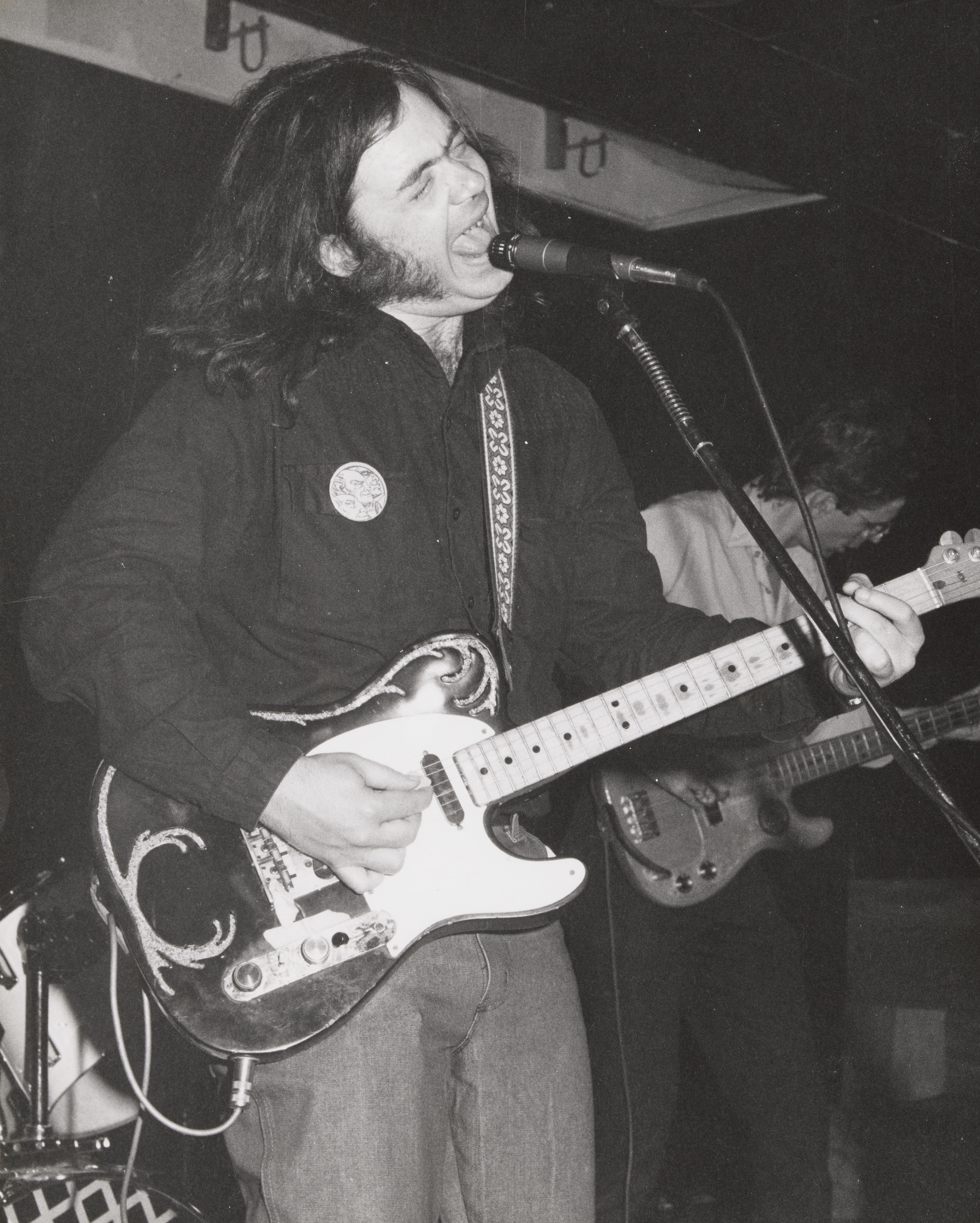 Roky Erickson in a black button-down shirt playing guitar and singing into a microphone on stage.